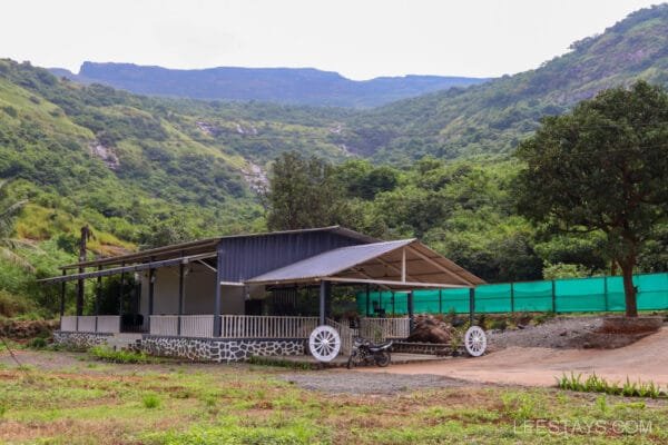 A cozy resort in Lonavala set amidst lush green hills, featuring a spacious building with a large porch and a scenic backdrop of mountains.