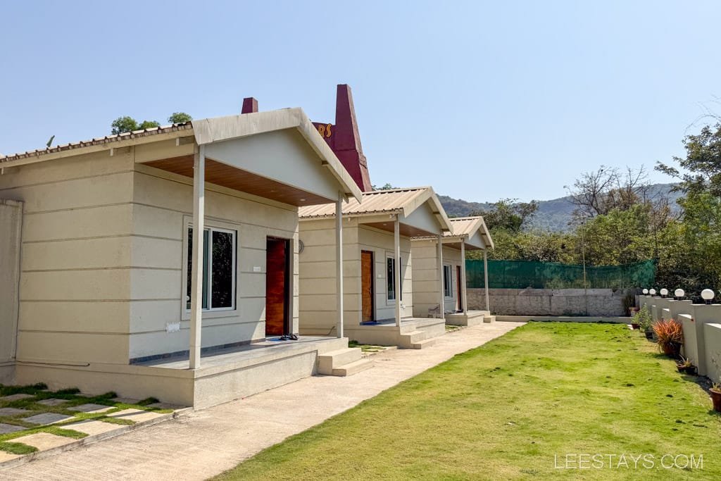 Row of quaint cottages in Lonavala, surrounded by greenery and hills, featuring a manicured lawn and a clear blue sky.