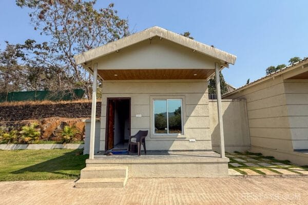 A small, beige cottage in Lonavala, surrounded by greenery and a clear blue sky overhead, with a chair on the front porch.