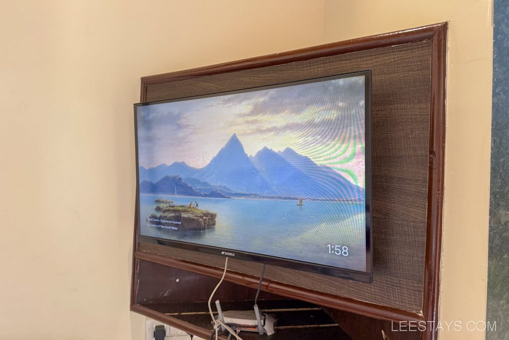 A wall-mounted TV in a cottage room in Lonavala, showing a serene mountain and lake landscape with the time displayed as 1:58.