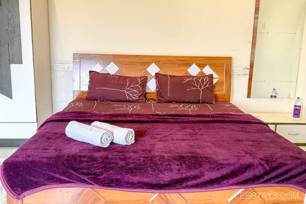 Cozy bedroom in a Lonavala cottage featuring a double bed with a plush maroon blanket and pillows, accompanied by two rolled towels.