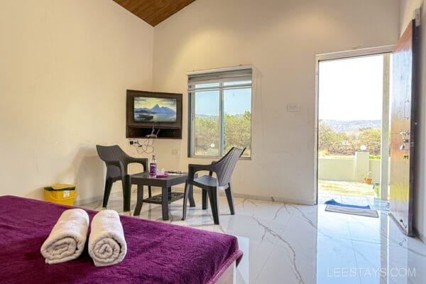 Cozy interior of a cottage in Lonavala, featuring a bed with folded towels, a table with chairs, a TV, and a window with a scenic view.