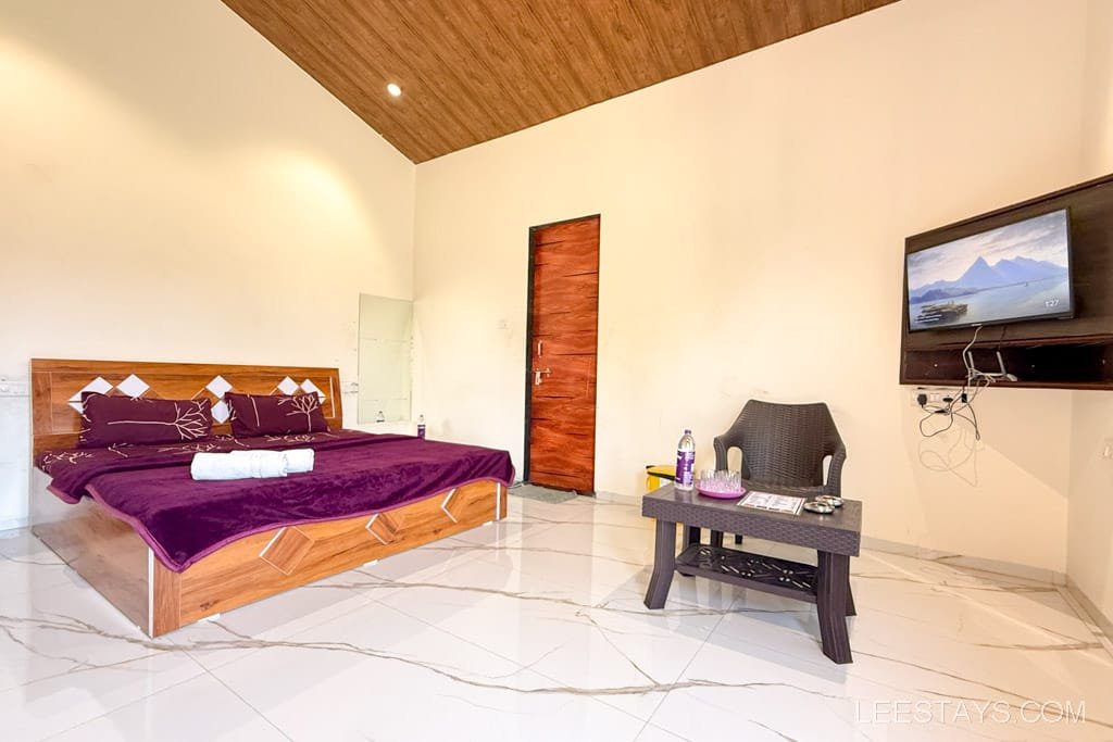 Interior of a cozy cottage in Lonavala featuring a bed with purple bedding, a small table with bottled water, and a mounted TV on the wall.